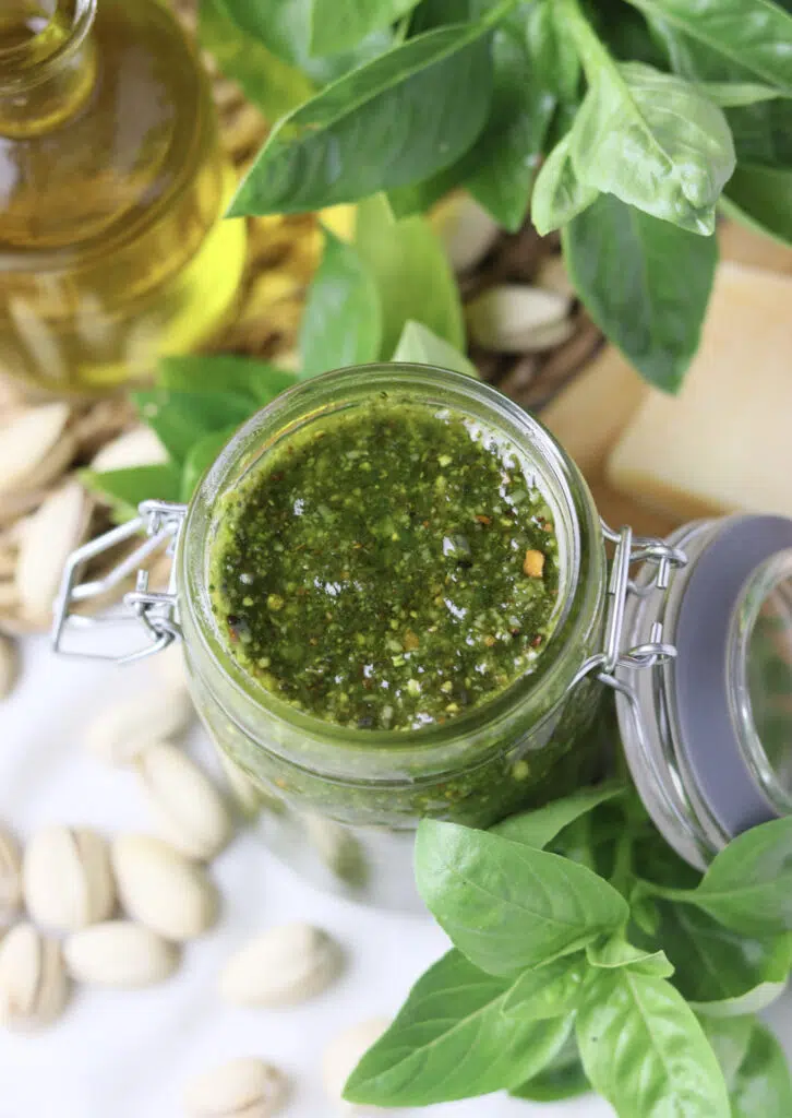 image showing basil pesto with pistachios in an opened jar with pistachios and olive oil in the background 