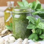 image of basil pesto with pistachios in a jar, with pistachios and olive oil in the background